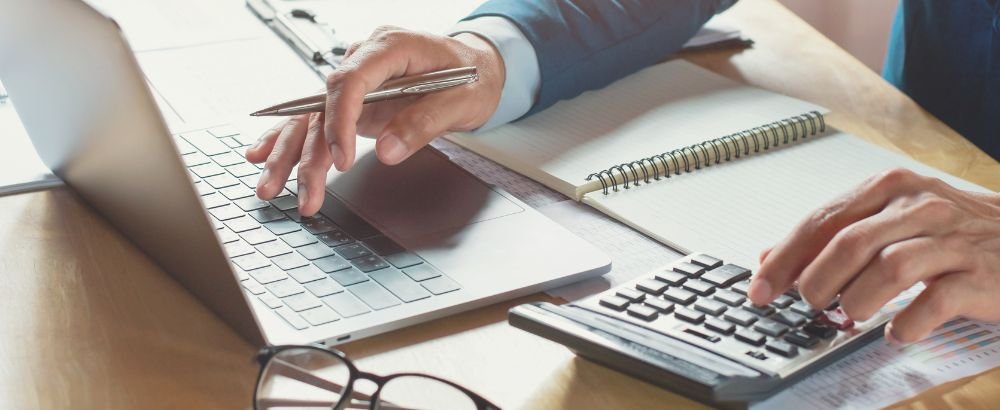 Man holding a pen working on the laptop while using calculator at the same time