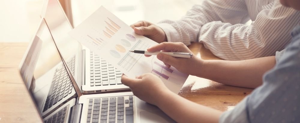 A laptop is kept on the table and two people are discussing and one person is holding a pen and another a chart paper