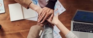 A diverse team's hands stacked together in a 'hands-in' gesture over a wooden desk.